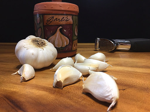 A few garlic cloves with a garlic press and a garlic keeper in the background.