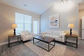 Model living room with cream couch and chair, two end tables with lamps, coffee table, patio door and window with blinds