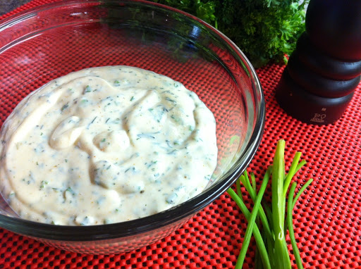 Ranch dressing in a glass bowl on a red table mat with green onions, parsley and a wood pepper mill.