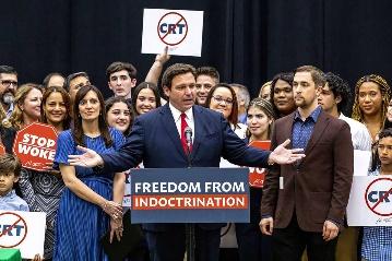 Florida Gov. Ron DeSantis, surrounded by supporters carrying signs saying Stop Woke and Ban CRT, holds out his hands wide at a podium marked: Freedom From Indoctrination.