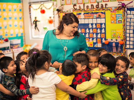 Mulher sorrindo em uma sala de aula cercada por um grupo de crianças.