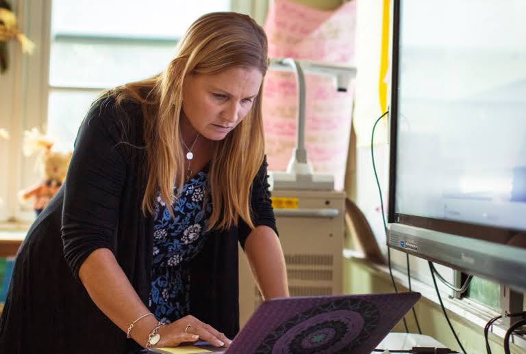 Uma educadora está em uma sala de aula trabalhando no laptop em uma mesa.